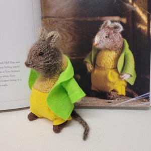 May include: Two knitted mouse figurines wearing yellow pants and green jackets. The mouse on the left is standing on a white surface, while the mouse on the right is standing on a wooden surface.