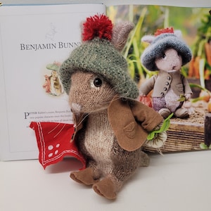 May include: A brown knitted rabbit wearing a green knitted hat with a red pom-pom, a brown jacket, and holding a red bandana. The rabbit is sitting on a white surface.