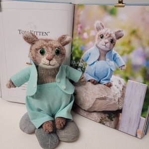 May include: Two knitted toy kittens wearing blue and green outfits. The kitten on the left is wearing a green jacket and pants. The kitten on the right is wearing a blue jacket and pants. Both kittens have brown fur and green eyes. The text "Tom Kitten™" is visible in the background.