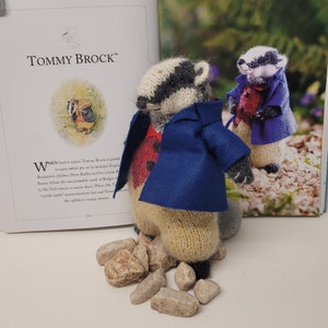 May include: A knitted badger toy wearing a blue jacket and red vest, standing on small gray rocks. The toy is based on the character Tommy Brock from the book "The Tale of Peter Rabbit".