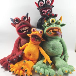 May include: A collection of four handmade knit monster dolls in red, orange, green, and dark grey. Each creature features distinct characteristics including multiple eyes, sharp teeth, horns, and clawed feet, crafted from textured knit fabric.