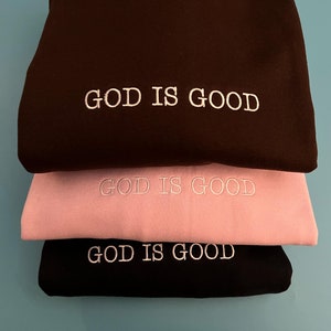 May include: Three sweatshirts stacked on top of each other. The top sweatshirt is black, the middle sweatshirt is pink, and the bottom sweatshirt is black. All three sweatshirts have the words "GOD IS GOOD" embroidered in white on the front.
