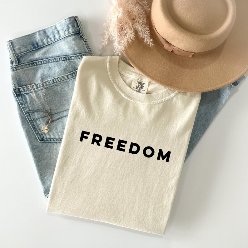May include: Cream-colored t-shirt with the word "FREEDOM" in black block letters. The shirt is paired with light blue denim jeans, a tan hat, and a delicate necklace. A sprig of dried flowers adds a touch of natural texture.