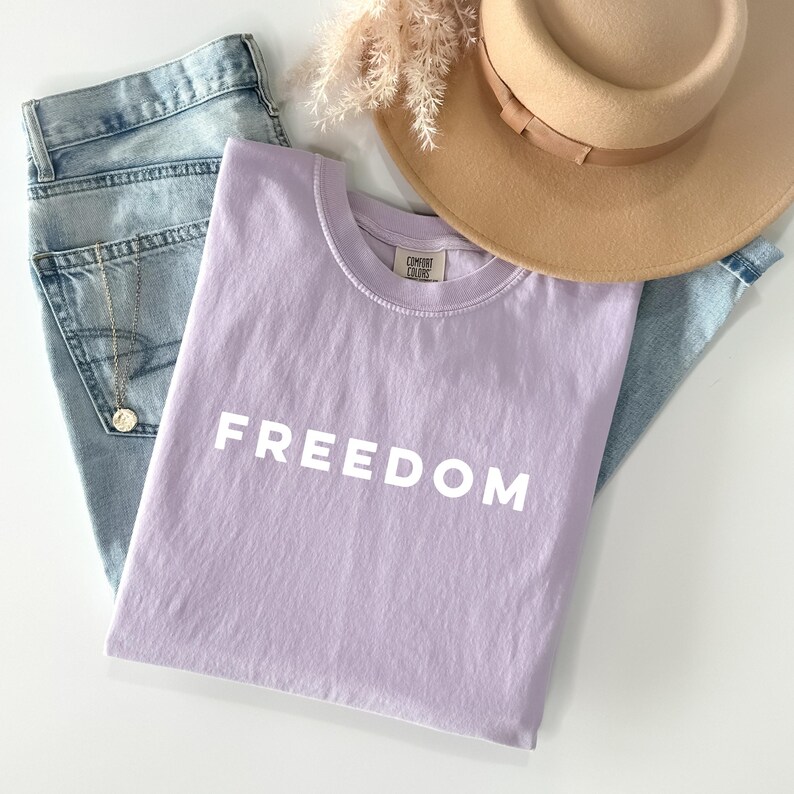 May include: A lilac-colored t-shirt with the word "FREEDOM" printed in white. The shirt is folded and placed on top of a pair of light blue jeans, a tan hat, and a gold necklace.
