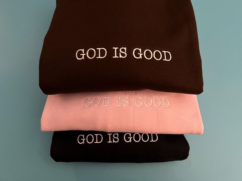 May include: Three sweatshirts stacked on top of each other. The top sweatshirt is black, the middle sweatshirt is pink, and the bottom sweatshirt is black. All three sweatshirts have the words "GOD IS GOOD" embroidered in white on the front.