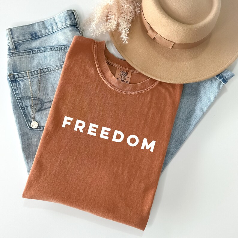 May include: A rust-colored t-shirt with the word "FREEDOM" in white block letters, laid on top of light blue denim jeans. A tan fedora hat and a gold necklace are also visible. The shirt is the focal point of this casual fashion ensemble.