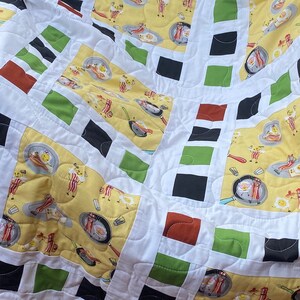 May include: A white quilt with a green, red, and black geometric pattern. The quilt features a repeating pattern of squares and rectangles. The fabric has a print of breakfast food, including eggs, bacon, and toast.