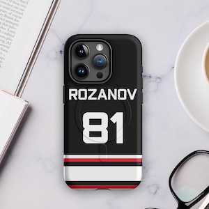 May include: Black phone case featuring the name "ROZANOV" and the number "81" in white. The case has a black, red, and white striped design at the bottom. The phone case is on a marble surface.