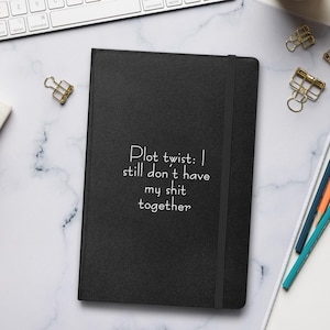 May include: A black notebook with the text "Plot twist: I still don't have my shit together" in white. The notebook is on a white surface with a keyboard, coffee cup, and pencils.