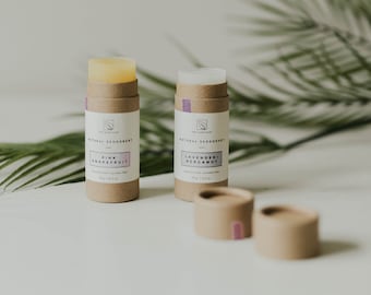 May include: Two natural deodorant sticks, one pink grapefruit and one lavender bergamot, on a white surface with a palm leaf in the background. The sticks are in cardboard tubes with the scent printed on the front.