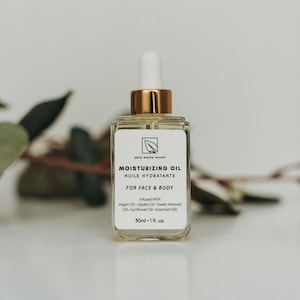 May include: A clear glass bottle with a gold lid and dropper, filled with a light yellow liquid. The bottle label reads "zero waste mvmt" and "Moisturizing Oil - Huile Hydratante" in black text. The label also states "For Face & Body" and lists the ingredients: "Infused With Argan Oil - Jojoba Oil - Sweet Almond Oil - Sunflower Oil - Essential Oils". The bottle contains 50 ml or 1 fl. oz. of oil.