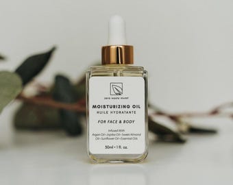 May include: A clear glass bottle with a gold lid and dropper, filled with a light yellow liquid. The bottle label reads "zero waste mvmt" and "Moisturizing Oil - Huile Hydratante" in black text. The label also states "For Face & Body" and lists the ingredients: "Infused With Argan Oil - Jojoba Oil - Sweet Almond Oil - Sunflower Oil - Essential Oils". The bottle contains 50 ml or 1 fl. oz. of oil.