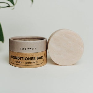 May include: A brown cardboard tube with the text "ZERO WASTE CONDITIONER BAR cedar + patchouli" printed on it. Next to the tube is a round, light pink conditioner bar.