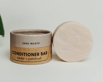 May include: A brown cardboard tube with the text "ZERO WASTE CONDITIONER BAR cedar + patchouli" printed on it. Next to the tube is a round, light pink conditioner bar.