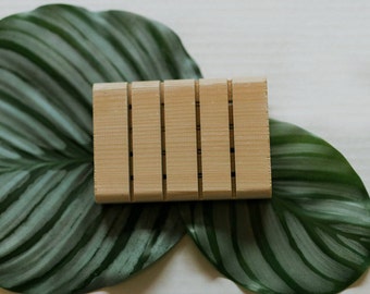 May include: A wooden soap dish with five slots for bar soap. The dish is sitting on a green leaf with white stripes.