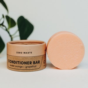 May include: A brown cardboard container with the text "ZERO WASTE CONDITIONER BAR sweet orange + grapefruit" on it. Next to the container is a pink conditioner bar.