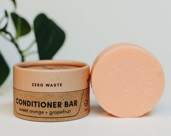 May include: A brown cardboard container with the text "ZERO WASTE CONDITIONER BAR sweet orange + grapefruit" on it. Next to the container is a pink conditioner bar.