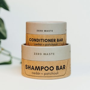 May include: Two brown cardboard containers with white text. The top container says "ZERO WASTE CONDITIONER BAR cedar + patchouli". The bottom container says "ZERO WASTE SHAMPOO BAR cedar + patchouli".