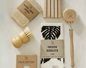 May include: Zero waste kitchen essentials set with a natural loofah sponge, a wooden soap dish, a dish brush, a bar of solid dish soap, and a Swedish dishcloth. The dishcloth is white with a black monstera leaf pattern and the text "Swedish Dishcloth, 2 Pack, Biodegradable, Plastic-Free, Reusable, Zero Waste Mvmt". The dish soap bar is labeled "Solid Dish Bar, Savon a Vaissele Solide, Bergamot Lime, Zero Waste Mvmt".