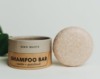 May include: A brown cardboard container with the text "ZERO WASTE SHAMPOO BAR cedar+patchouli" on it. Next to the container is a round, light brown shampoo bar.