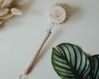 May include: A wooden dish brush with a natural bristle head and a wooden handle. The brush has a round wooden head with the text "zero waste mvmt" and a leaf logo engraved on it.