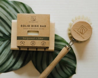 May include: A brown cardboard box with the text "SOLID DISH BAR" and "bergamot + lime" on it. The box is sitting on a wooden soap dish. A wooden brush with a white bristle head is next to the box. The brush has a metal loop and the text "zero waste mvmt" on the head.