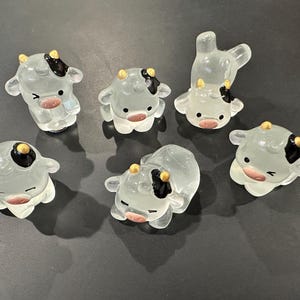 May include: Six small glass figurines of cows in various poses. The cows are white with black spots and gold accents. They are all facing forward.