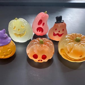 May include: A collection of six Halloween-themed pumpkin figurines in various shapes and colors. The pumpkins are orange, pink, and yellow, with carved faces and some with hats. The figurines are arranged on a dark surface.