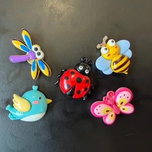 May include: A collection of five colorful insect and animal pins including a blue and yellow dragonfly, a yellow and black striped bee, a red and black ladybug, a blue and yellow bird, and a pink and yellow butterfly with detailed patterns.