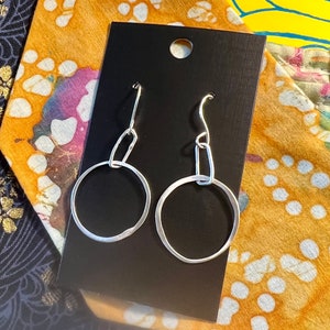 May include: A pair of silver hoop earrings with a simple, minimalist design. The earrings are displayed on a black card with a white circle cutout.