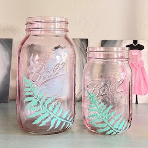 May include: Two pink Ball mason jars, one large and one small, with a fern design in turquoise. The jars are clear and have a vintage aesthetic. The Ball logo is visible on the front of each jar.