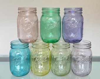 May include: A collection of seven vintage Ball Mason jars in various pastel colors, including pink, green, blue, yellow, and purple. The jars are arranged in a stacked formation, showcasing their classic design and embossed lettering.