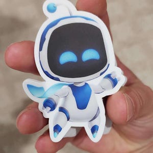 Peut inclure: Un autocollant blanc et bleu représentant un personnage de robot de dessin animé avec un visage noir et des yeux bleus. Le robot a une cape bleue et tend les bras. L'autocollant est tenu dans la main d'une personne.