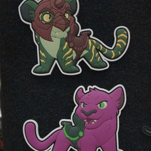 May include: Two cartoon animal patches, one green and yellow with a red helmet and the other purple with a green helmet. Both patches have a white outline.