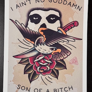 Op de afbeelding: Een vintage-stijl tattoo-flashprint met een zwart-witte schedel met een havik die erboven vliegt en een dolk in zijn klauwen vasthoudt. De havik zit op een rode roos met groene bladeren. De print heeft een bruine achtergrond met een vervaagde look en de tekst "I ain't no goddamn son of a bitch".