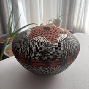 May include: A round, low-profile ceramic vessel featuring a small circular opening at the top. The surface is decorated with intricate black and white cross-hatched geometric patterns, accented by terracotta-colored bands and a textured top section.