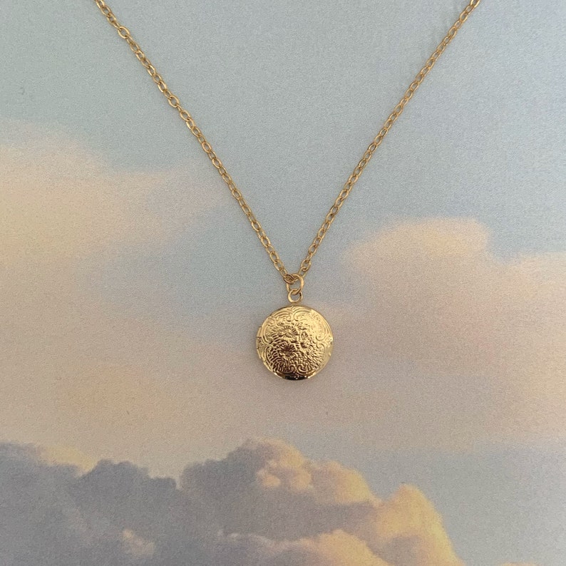 May include: A gold-colored necklace with a round pendant. The pendant features an ornate, embossed floral design. The chain is delicate and the background is a soft, cloudy sky.