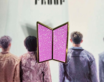 May include: A pink and gold pin featuring a stylized open book design with a glittery texture. The pin is set against a background with the word "PROOF" in black letters and images of people.