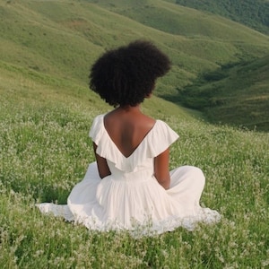 May include: A person wearing a white dress with a low V-neck and ruffled sleeves sits in a field of green grass and small white flowers. The background features rolling green hills under a bright sky.