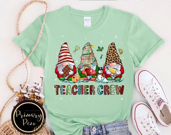 May include: A light green t-shirt featuring a graphic of three gnomes with different hats, holding school supplies like a book, apple, and pencil. The text "TEACHER CREW" is printed below the gnomes in colourful, patterned block letters.