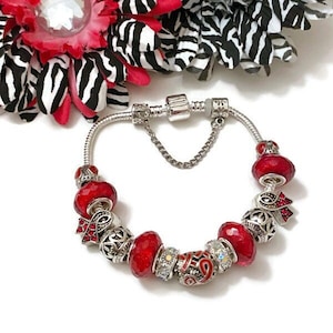 May include: A silver charm bracelet with red glass beads and a red ribbon charm. The bracelet has a safety chain and a lobster clasp.