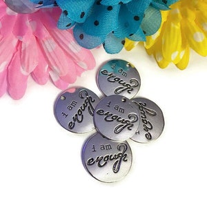 May include: Five silver metal charms with the words "I am enough" written on them. The charms are arranged in a pile on a white background. The charms are surrounded by pink, blue, and yellow flowers.
