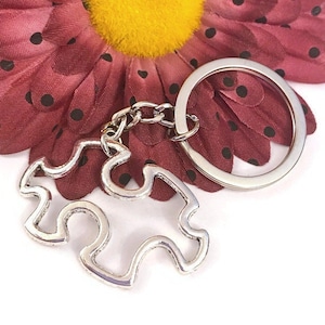 May include: A silver-toned puzzle piece keychain with a silver chain and a silver ring. The keychain is lying on a white surface with a red flower with black polka dots in the background.