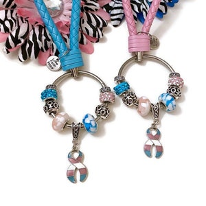 Könnte beinhalten: Zwei Schlüsselanhänger mit einem silbernen Ring und Perlen. Ein Schlüsselanhänger hat ein hellblaues geflochtenes Lederband und einen hellblauen, rosa und weißen Bandanhänger. Der andere Schlüsselanhänger hat ein rosa geflochtenes Lederband und einen hellblauen, rosa und weißen Bandanhänger. Beide Anhänger haben den Text "Love Faith Hope" auf einem kleinen silbernen Anhänger.