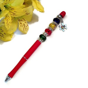 May include: Red pen with a silver top and a charm featuring a puzzle piece design. The charm is attached to a string of colourful beads, including red, green, yellow, and blue.