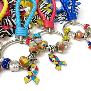 May include: Five colorful keychains with a silver ring and charms. Each keychain has a different colored braided strap. The charms include a puzzle piece, a ribbon, and colorful beads. The charms are attached to the ring with a silver chain.