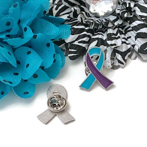 Puede incluir: Dos pines de cinta azul verdoso y morado. Los pines están sobre un fondo blanco. Un pin es plateado y el otro es azul verdoso y morado.