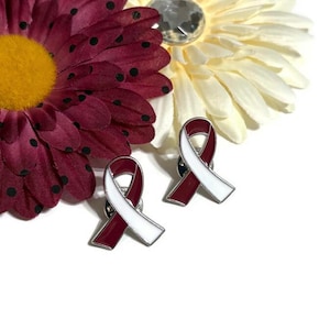 May include: Two maroon and white enamel ribbon pins. The ribbons are tied in a bow shape and have a silver metal backing.