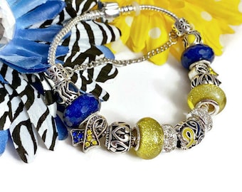 May include: A silver charm bracelet with blue and yellow beads, including a charm with a yellow and blue ribbon. The bracelet is on a white background.