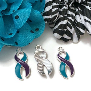 Puede incluir: Tres colgantes de cinta de concienciación. Dos son de esmalte turquesa y morado, y uno es plateado. Los colgantes tienen forma de cinta, con un lazo en la parte superior para sujetarlos a un collar o pulsera. El fondo incluye una flor turquesa y una flor con estampado de cebra en blanco y negro.